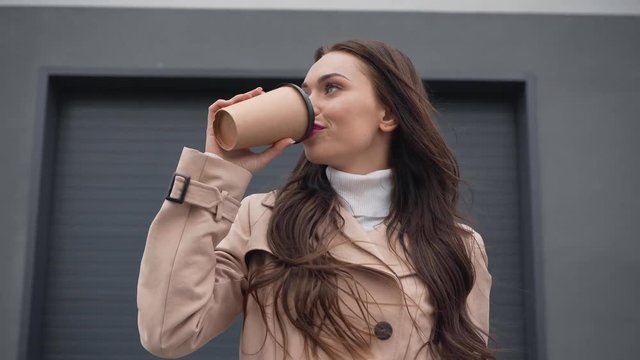 smiling elegant woman in beige trench coat walking and drinking coffee from paper cup in windy weather