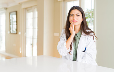 Young woman wearing medical coat at the clinic as therapist or doctor touching mouth with hand with painful expression because of toothache or dental illness on teeth. Dentist concept.
