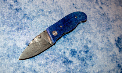 Damascus pocket knife with blue camel bone handle displayed on blue and white fabric.