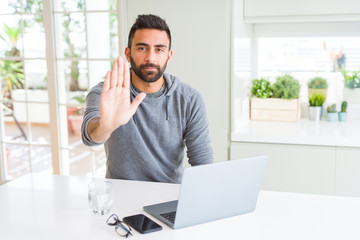 Handsome hispanic man working using computer laptop with open hand doing stop sign with serious and confident expression, defense gesture