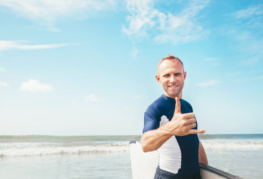 Young man Surfer portrait showing surfer's famous Shaka sign gesture in camera when he comming with long surf board to waves. Active holidays spending concept.