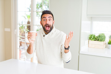 Handsome hispanic man drinking a coffee in a paper cup very happy and excited, winner expression celebrating victory screaming with big smile and raised hands
