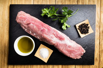 pork tenderloin with salt, black pepper, a bowl of oil and parsley on a black slate plate
