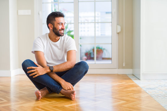 Handsome hispanic man wearing casual t-shirt sitting on the floor at home looking away to side with smile on face, natural expression. Laughing confident.
