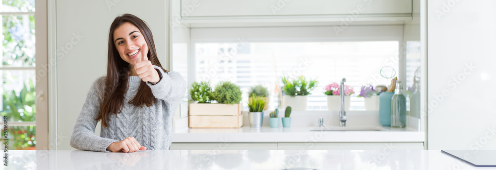 Poster Wide angle picture of beautiful young woman sitting on white table at home doing happy thumbs up gesture with hand. Approving expression looking at the camera with showing success.