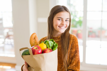 Beautiful young girl holding paper bag of fresh groceries with a happy face standing and smiling with a confident smile showing teeth