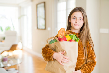 Beautiful young girl holding paper bag of fresh groceries with a confident expression on smart face thinking serious