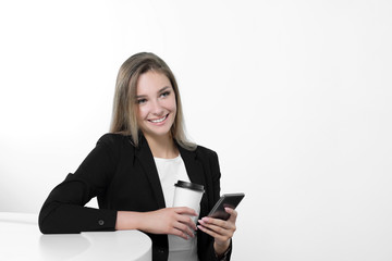 A young beautiful woman in a black suit and white blouse is sitting at the office table. Drinks a hot drink of coffee from a white paper cup. Writes a message on a smartphone, communicates and smiles.