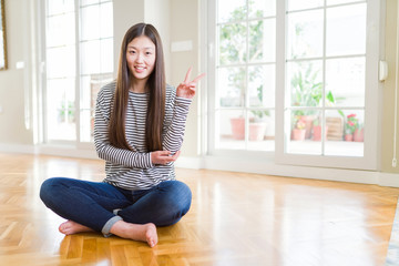 Beautiful Asian woman sitting barefoot on the floor at home smiling with happy face winking at the camera doing victory sign. Number two.
