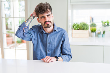 Fototapeta premium Young man wearing casual shirt sitting on white table confuse and wonder about question. Uncertain with doubt, thinking with hand on head. Pensive concept.
