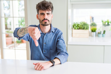 Fototapeta premium Young man wearing casual shirt sitting on white table looking unhappy and angry showing rejection and negative with thumbs down gesture. Bad expression.