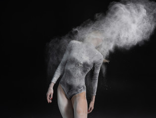 Beautiful slim girl wearing a black gymnastic bodysuit covered with white powder and dust flies from her hair on a dark. Artistic conceptual photo.