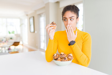 Young african american woman eating chocolate chips muffins cover mouth with hand shocked with shame for mistake, expression of fear, scared in silence, secret concept