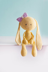 Soft toy knitted hare. Sitting yellow sad plush hare with a violet bow. Amigurumi.