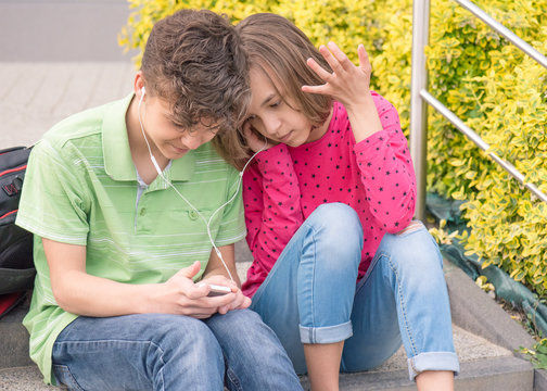 Happy teenage boy and girl with headphones are using gadget, talking and smiling while sitting on the stairs outdoors. Young sister and brother teens playing on mobile phone and listening to music.