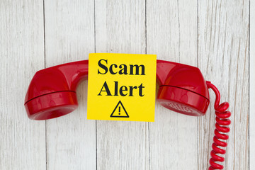 Scam alert message on sticky note on retro red phone on weathered whitewash textured wood background