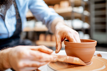 Man making clay jug forming shape by hands on the pottery wheel indoors, close-up view