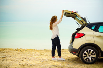 Happy stylish young woman traveler on beach road near white crossover car, holding hat in hand. Banner. Travel, summer vacation, holiday, freedom concept. Digital detox