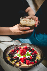 a young woman sits comfortably in an armchair in the morning sun, eats granola and drinks a coffee with whipped cream and cinnamon