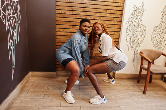 Two african womans in stylish casual clothes posing against wooden wall sat down and dance twerk.