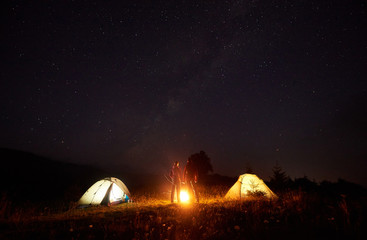 Night camping picture. Bright bonfire burning between two silhouettes of boy and girl standing in front of illulminated tents, holding hands under beautiful dark starry sky on distant hills background