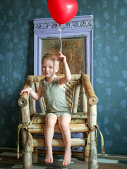 Fototapeta premium Funny boy in blue clothes and glasses is playing with red balloon against gray wall against background of ancient mirror. child is happy at home