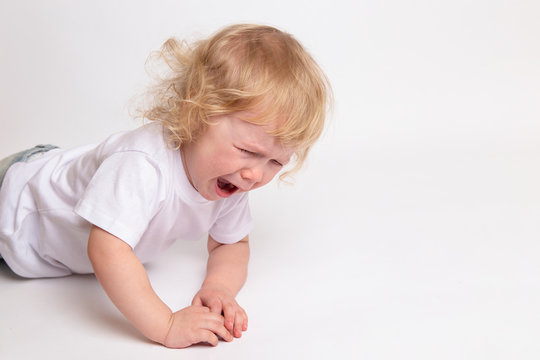 A little curly crying child in a clean white T-shirt is lying on the floor and crying.