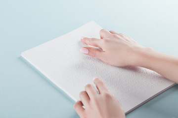 cropped view of young woman reading braille text on paper isolated on blue