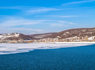Fototapeta premium Beautiful winter landscape with small European village on the side of a frozen lake. Winter vacation and holidays, New Year and Christmas recreation and relaxing. Resort, hotels.
