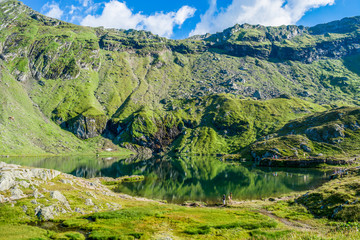 Fototapeta na wymiar Balea lake near Transfagarasan road in the Carpathian Mountains, Romania, Eastern Europe. Green meadows and water in the wilderness on a summer day. Vacation and travel destination. 
