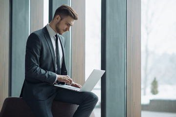 Fototapeta premium Young handsome man sitting in office with cup of coffee and working on project connected with modern cyber technologies. Businessman with notebook trying to keep deadline in digital marketing sphere.