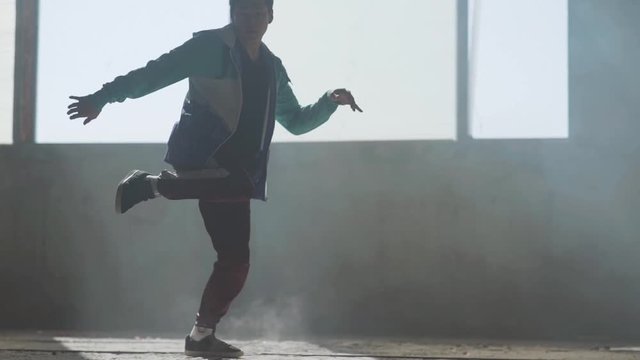 Experienced young hip-hop street dancer performing in front of large window in the dark abandoned building. The man making moves, jumping and crouches dancing.. Slow motion