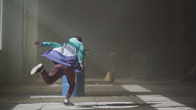 Young asian hip-hop male dancer in sport wear dancing in the dark abandoned building in front of blue gas barrel. Contemporary. Hip hop culture. Rehearsal. Slow motion.