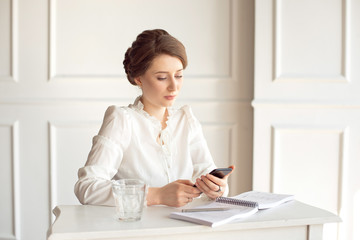 Portrait of a beautiful smiling young brunette businesswoman in a white shirt sitting on a bright modern workstation working with papers .