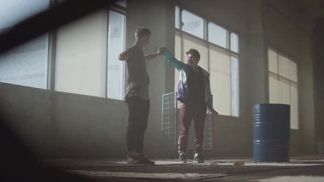 Two young men dancing in the dark and dusty room of abandoned building. Teenagers making dance move simultaneously, holding hands. Flexible men making wave with their bodies. Slow motion