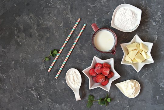 Ingredients for the preparation of a dessert or drink : white chocolate, frozen strawberry, powdered sugar, milk, corn starch, fresh mint and whipped cream for decoration, straws. Top view, copy space