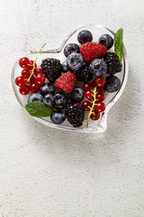 Fresh ripe berries in a glass heart-shaped bowl on a stone white table. summer season. healthy recipes and drinks