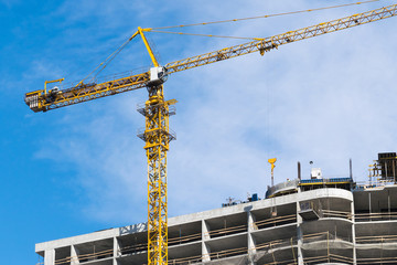 high-rise construction crane with a long arrow of yellow color against the blue sky over a new multi-storey building of concrete and brick under construction