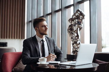 Young handsome man sitting in office with cup of coffee and working on project connected with modern cyber technologies. Businessman with notebook trying to keep deadline in digital marketing sphere.