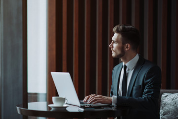 Young handsome man sitting in office with cup of coffee and working on project connected with modern cyber technologies. Businessman with notebook trying to keep deadline in digital marketing sphere.