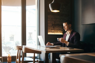 Young handsome man sitting in office with cup of coffee and working on project connected with modern cyber technologies. Businessman with notebook trying to keep deadline in digital marketing sphere.