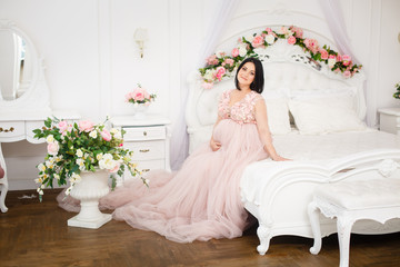 Fototapeta na wymiar Young pregnant woman in a light classic interior. Caucasian woman with black hairs in long romantic dress near the large window. Concept of new life. Waiting of baby. Pretty pregnant woman in a room