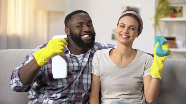 Happy mixed-race couple in gloves with cleanser spray and sponge, householding