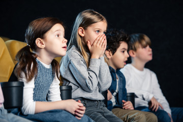 cute child holding hands on face while watching movie with multicultural friends