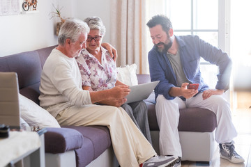 Adult and senior family at home with parents and son - caucasian people sitting on the sofa working with technology devices laptop and phone together enjoying internet connection