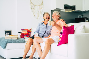 Happy old senior caucasian couple man and woman together at home sitting on the sofa drinking...