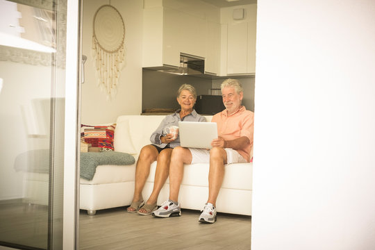 Elderly lifestyle with caucasian old aged mature couple at home sitting on the white sofa and enjoying technology laptop together in relax - senior people and computer concept