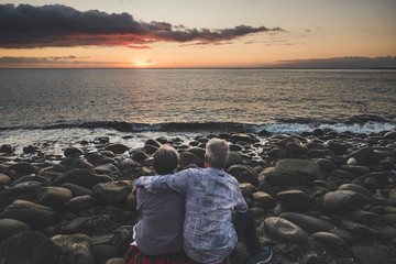 Senior couple viewed from back hug and love enjoying the sunset at the berach during summer...