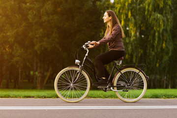 Naklejka na ściany i meble Young attractive woman riding through the park after work. Beautiful lady cycling during sunset. Bike as a trendy transport. Healthy outdoors activity on a warm summer day. Bicycle trend in the city.