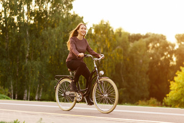 Obraz na płótnie Canvas Young attractive woman riding through the park after work. Beautiful lady cycling during sunset. Bike as a trendy transport. Healthy outdoors activity on a warm summer day. Bicycle trend in the city.
